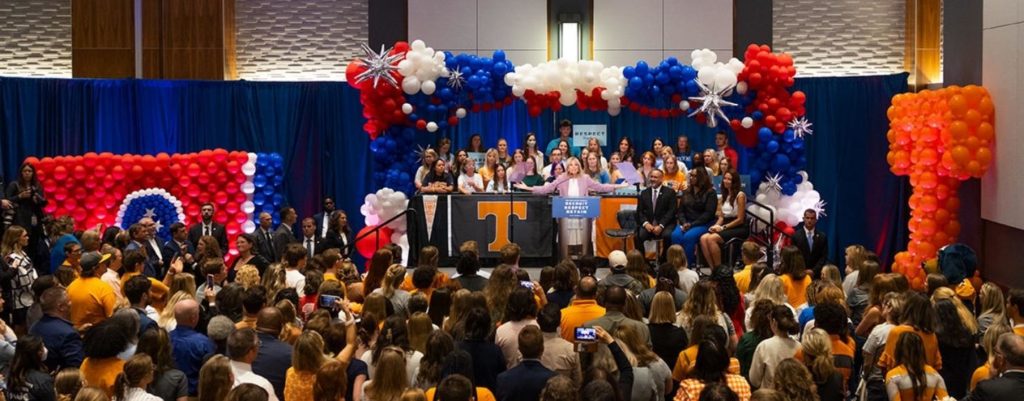 First Lady Jill Biden standing at a podium with arms outstretched in a room full of people.