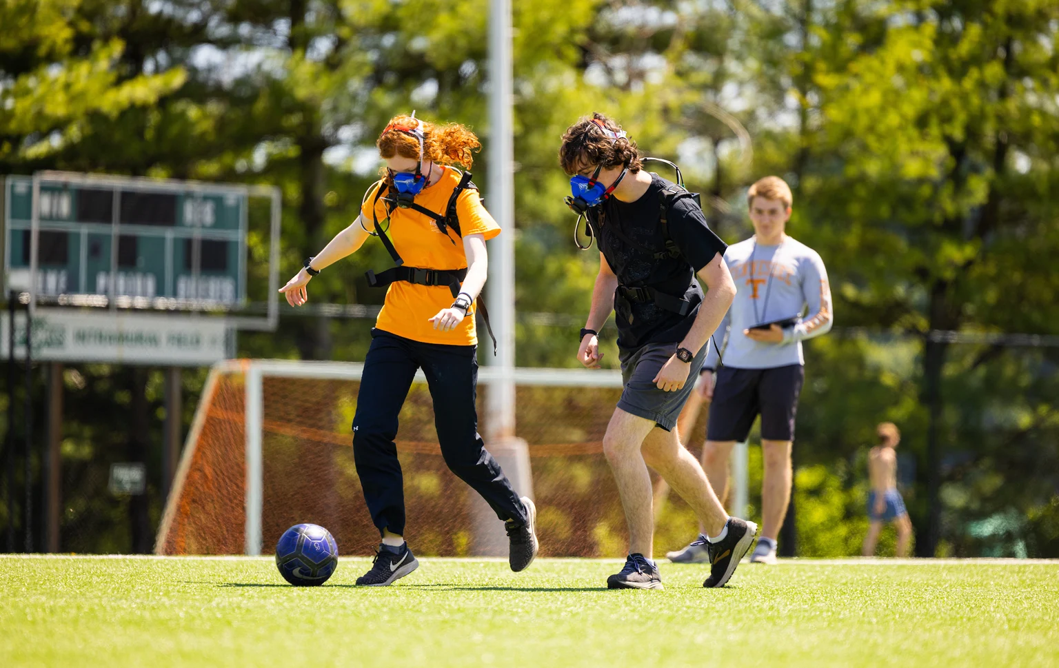 Students in a field soccer game wearing portable metabolic masks while a researcher observes