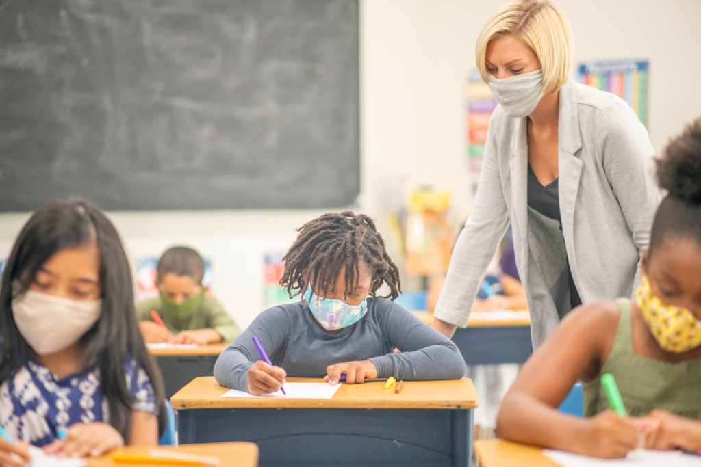 White female teacher inside multi-racial classroom assisting elementary students with assignment.