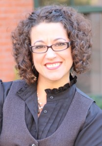 Jennifer Jabson. A feminine presenting person with tan skin smiles at the camera. She has dark curly brown hair to her shoulders and dark framed glasses. She wears a high collar black shirt with a black vest.