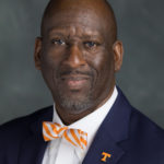 Portrait photo of Dorian McCoy. A bald Black man with dark skin smiles at the camera wearing a navy suit jacket, white shirt, and checkerboard bow tie.