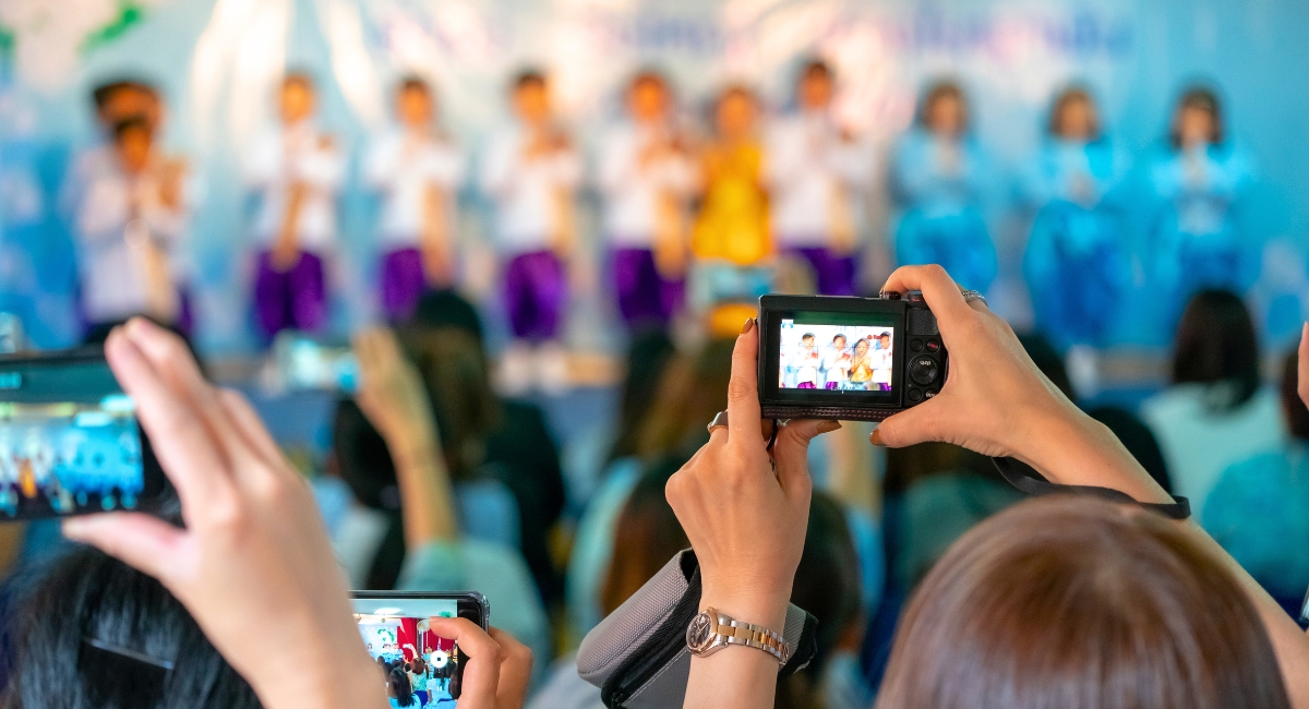 Parents sitting in the audience of a performance holding up digital cameras and smart phones to take pictures of children on a stage.