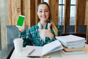 high school female giving thumbs up holding cell phone sitting at desk with books and coffee