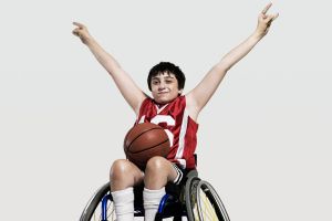 young boy wearing team basketball uniform and holding arms up with basketball on lap and sitting in adapted wheelchair