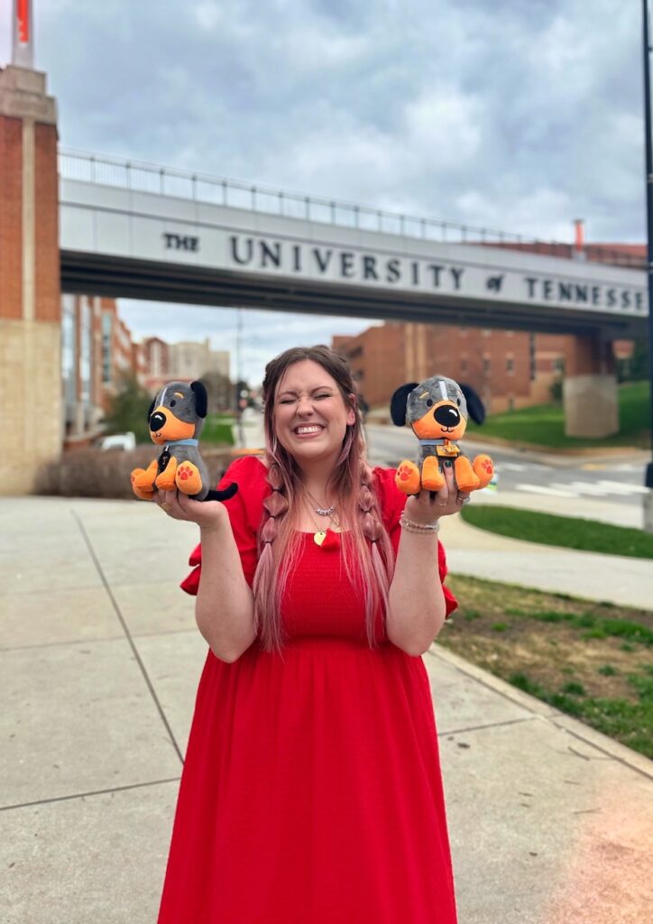 Rebekah Goode standing in front of the UT Pedestrian Bridge smiling with her eyes closed and holding a Baby Smokey plush in each hand.