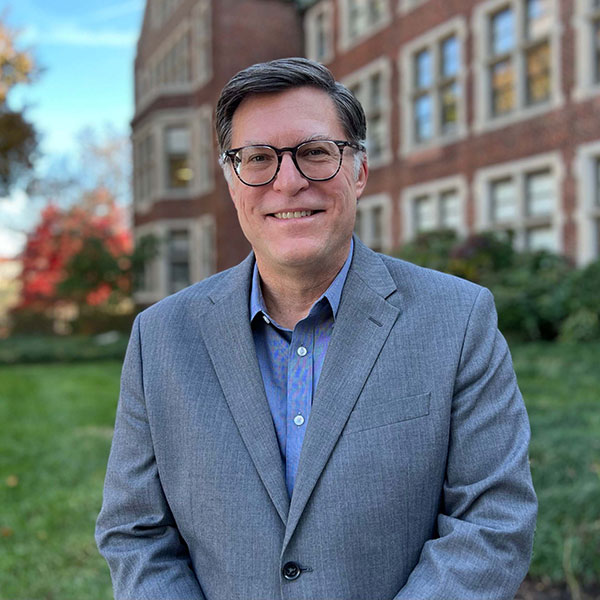 Doug Edlund, Director CEHHS Marketing and Communications. A white man stands in front of a campus building smiling at the camera. He has short dark brown hair and glasses. He is wearing a grey blazer and light blue shirt.