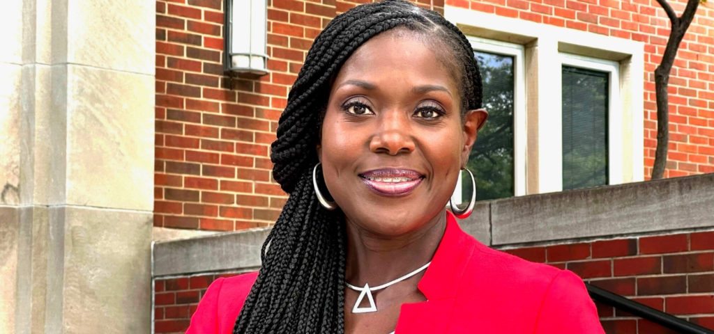 Kimberly R. Hill smiles at the camera. She is a Black woman with long braided hair pulled to one side and is wearing a red blazer and silver jewelry.