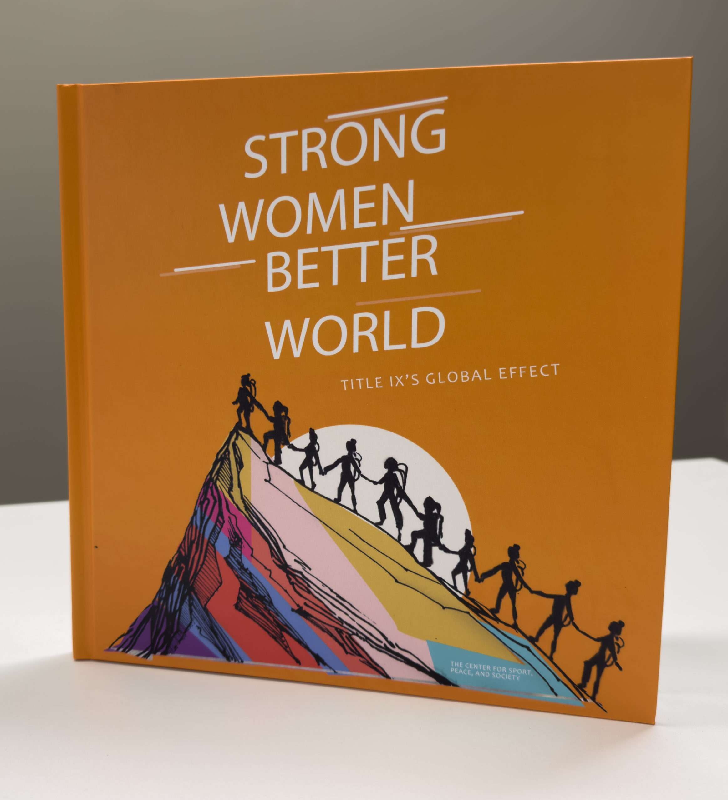 Photo of the book Strong Women Better World. The has an orange cover with the text, Strong Women Better World and has a drawing depicting a group of women silhouettes scaling a mountain. The book is placed on a table .