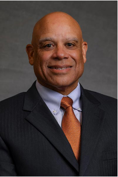 Portrait of Mark Dean. He has dark skin and is bald. He is wearing a black suit jacket, blue shirt, and orange necktie. He is smiling in the photo