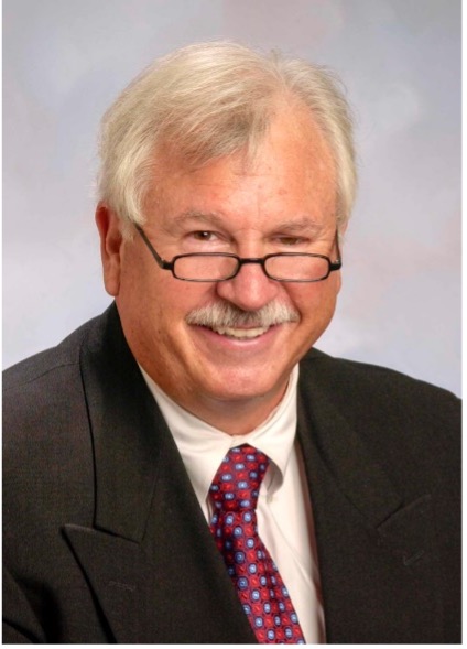 Portrait photo of Steve McCallum. He has fair skin, grey hair and a grey mustache. He is wearing plastic frame reading glasses, a dark suit jacket, white shirt and red necktie. He is smiling in the photo.