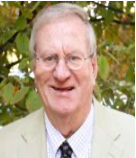 Portrait photo of S. Wayne Mulkey. He has fair skin, grey hair, and is wearing wire frame glasses. He is wearing a tan suit jacket, white shirt and red necktie. He is smiling in the photo