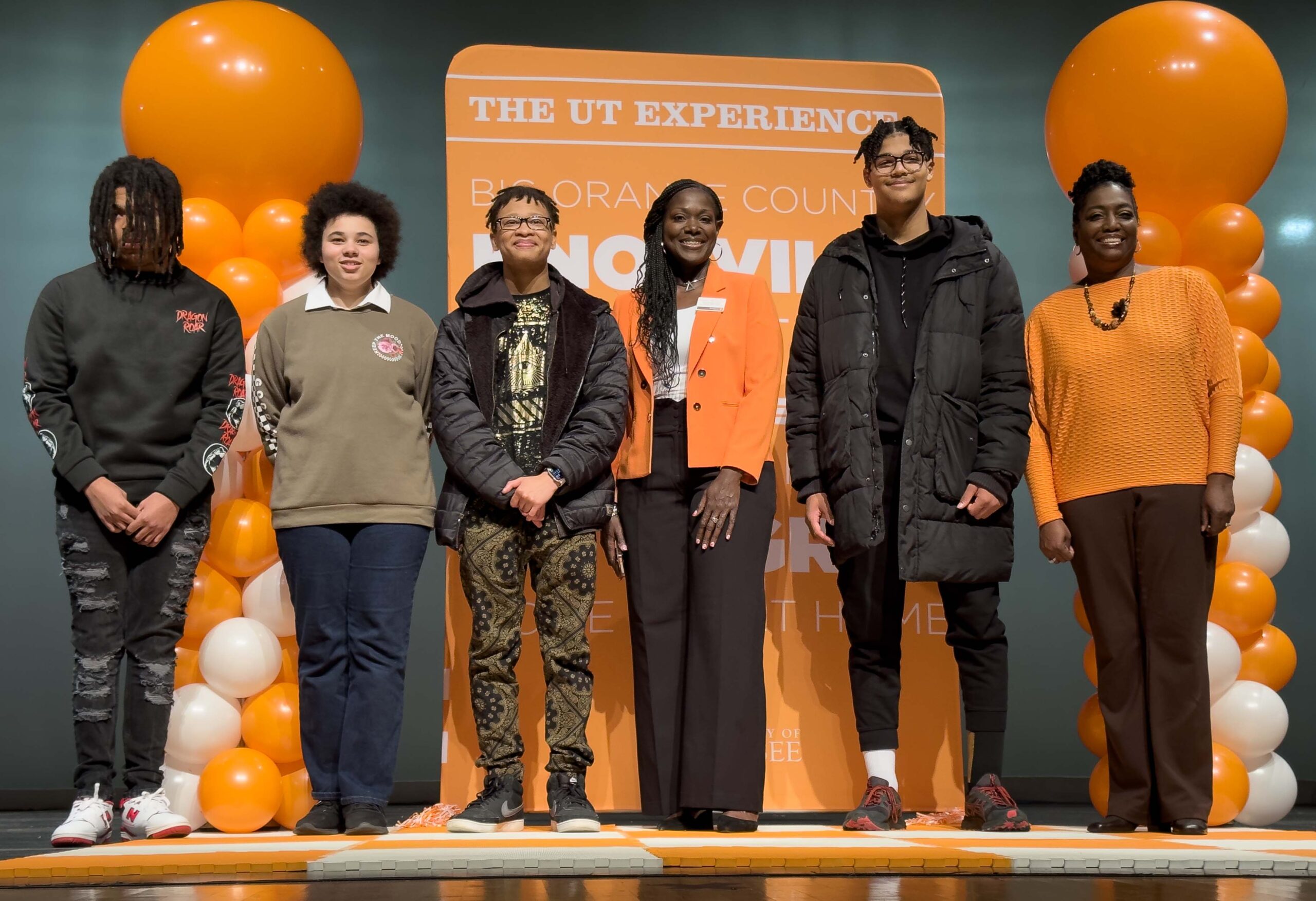 A group of Project Excellence students poses for a picture in front of an orange background with orange balloons on each side. There are three male and one non-binary students in the picture, along with Kimberly Hill and a teacher. All persons in the photo have dark skin and dark hair.