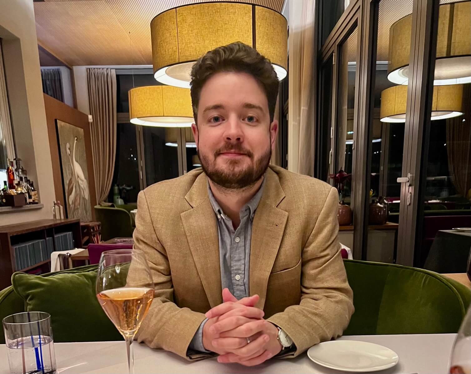 Jack Babb sits at a table in a restaurant setting. His forearms and hands are on the table. He has fair skin, dark brown hair, a beard and mustache. He is wearing a light tan sport coat and light grey shirt. 