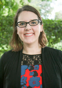 Ashlee ANDERSON. A feminine presenting White person with brown, shoulder-length hair and dark frame glasses. She wears a black blazer and a top with blue and red flowers.