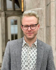 Patrick GRZANKA. A masculine presenting white person with short, straight icy-blond hair and dark glasses. He has a grey blazer and a white shirt with graphic icons.