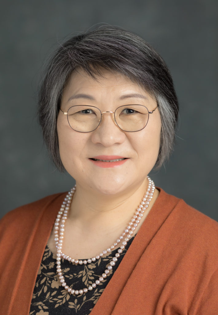 Portrait photo of Junhee Kwon, she is of Asian descent and has short dark hair . She is wearing glasses, a dark orange sweater and dark shirt. She is smiling in the picture.
