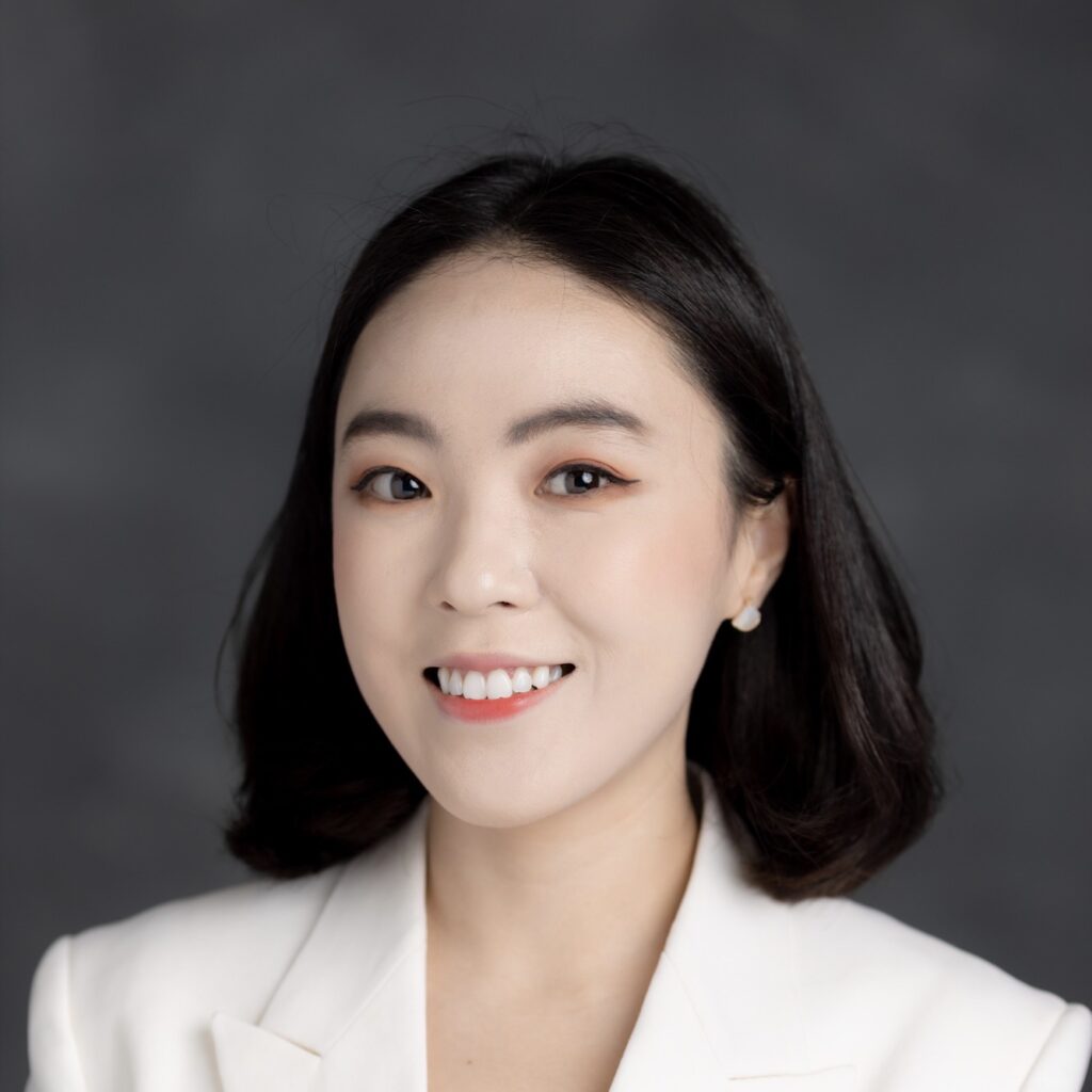Portrait photo of Dan Jin, an Asian female with dark hair. She is wearing a cream colored blazer and is posed in front of a dark gray background.