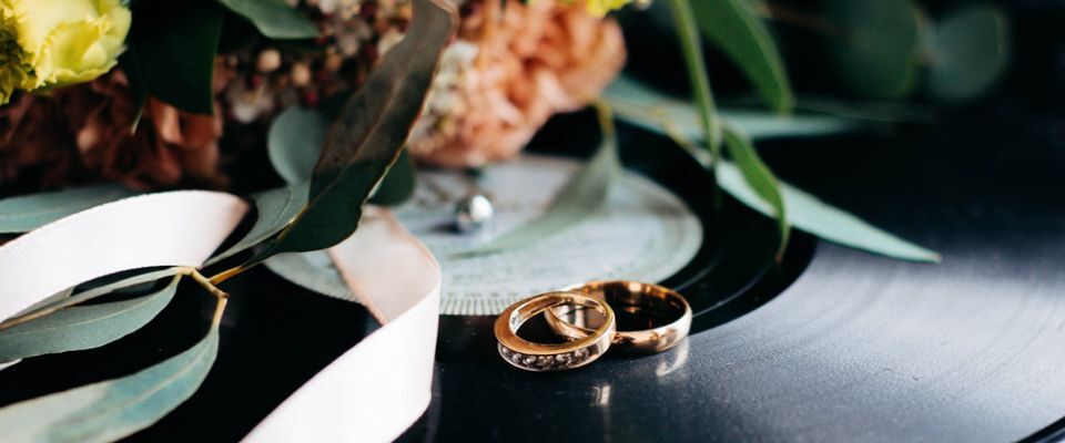 Photo illustration of two gold wedding rings overlapping. They are placed on a vinyl record along with a bouquet of flowers. 