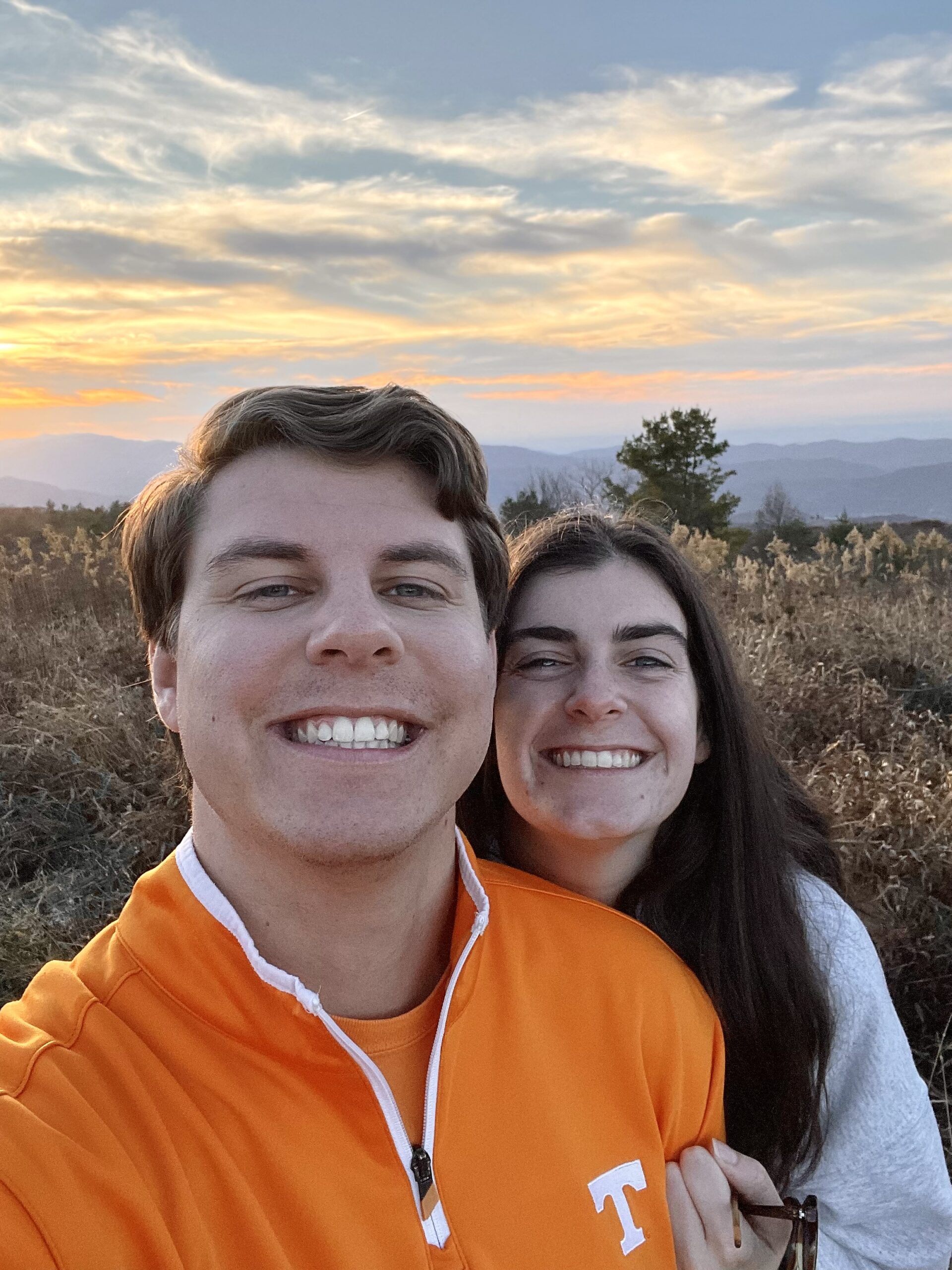 Photo of Jackson and Shelby. Both are fair skinned and have brown hair. Jackson is wearing an orange UT jacket, while Shelby is wearing a white jacket. They are posing on a mountain top with mountains in the background.