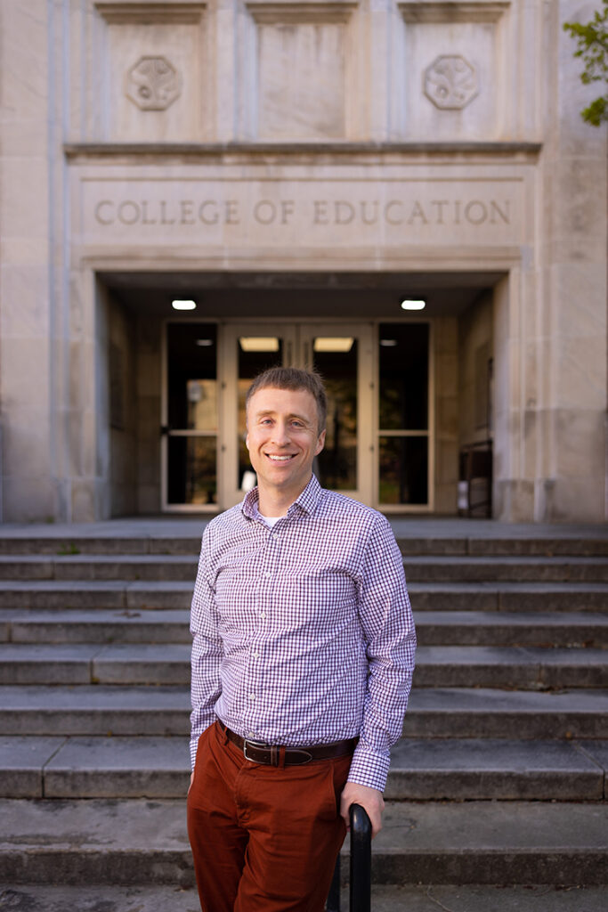 A portrait of Robert Kelchen in front of the Claxton Education Building. Kelchen has fair skin, short, light brown hair, and is wearing a burgundy and white plaid shirt and burgundy pants.