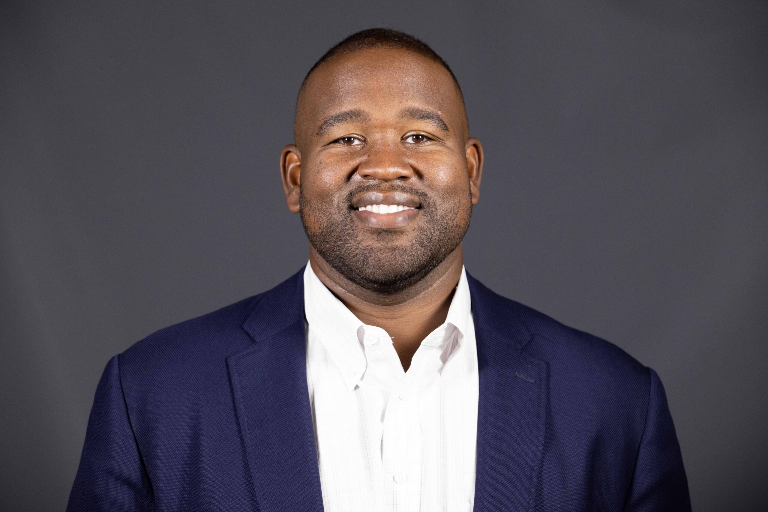 Portrait photo of Jacques McClendon. He is standing in front of a dark background. He has dark skin and short black hair. He is smiling in the photo