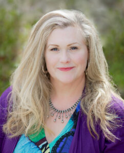 An outdoor portrait photo of Dr. Jillian C. Shipherd. She has fair skin, long blonde hair, and is wearing a colorful blouse and cardigan.