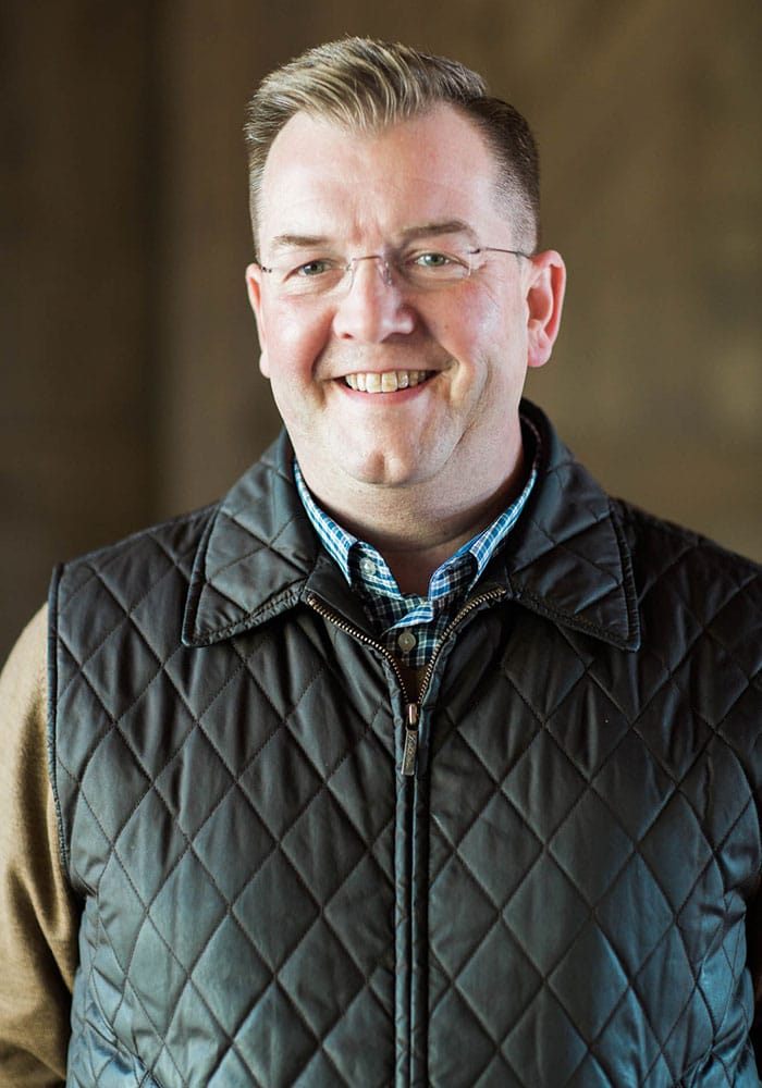 Portrait photo of Brian Lee. He has fair skin and short brown hair. He is wearing glasses, a green shirt and green vest. He is smiling in the photo.