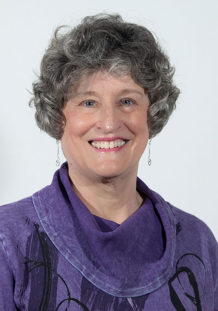 Photo of Linda Lee. She has fair skin, medium length grey hair and is wearing a purple top. She is smiling in the photo.