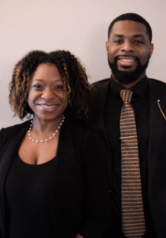 Photo of Monica and Chima Onwuka. They both have dark skin and dark hair. Monica is on the left and wearing a black top. Onwuka is on the right and wearing a black shirt and striped tie. Both are smiling in the photo