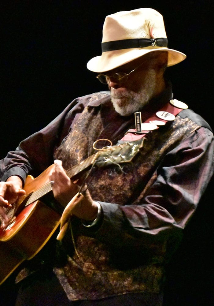 Photo of James "Sparky" Rucker playing a guitar on stage. He has dark skin and grey hair. He is wearing a hat and a dark shirt in the photo