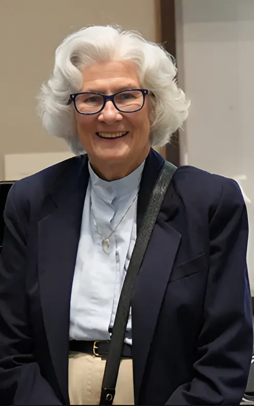 Portrait of Charmaine Mamantov—a woman with light skin, black glasses, and chin-length curled white hair—wearing a pale blue button-down shirt and a navy blazer, while standing in a beige room.