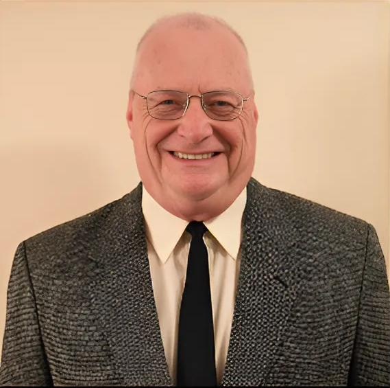 A portrait of David Royse—a man with light skin, simple silver metal framed glasses, and sparse gray and white hair on his head—wearing a white button-down shirt, black tie, and gray tweed blazer while standing against a cream wall.