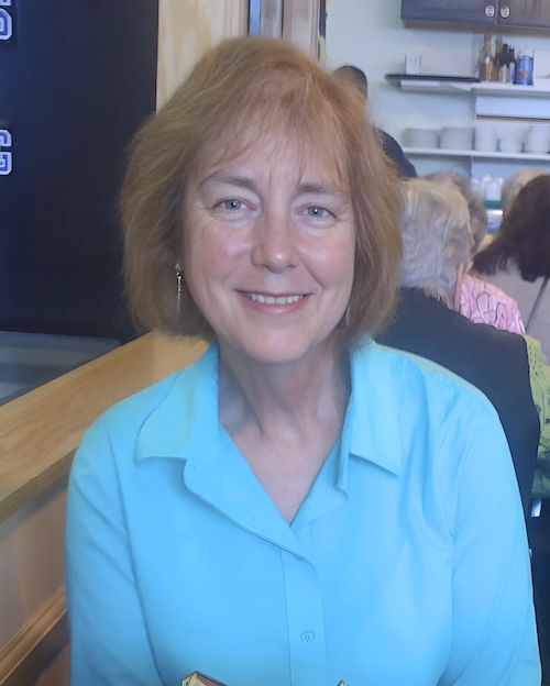 Portrait of Sarah DeYoung—a woman with light skin and chin-length light brown hair—smiling while sitting in a restaurant, wearing a light blue button-down shirt, with a warm, professional expression.