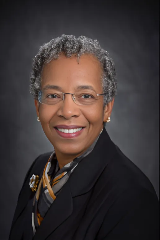 A portrait of Saundra McGuire—a woman with warm, brown skin, oval-shaped glasses without any frames around the lenses, and a short, curly gray pixie cut—while wearing a deep hued blouse and black blazer against a simple dark gray background.