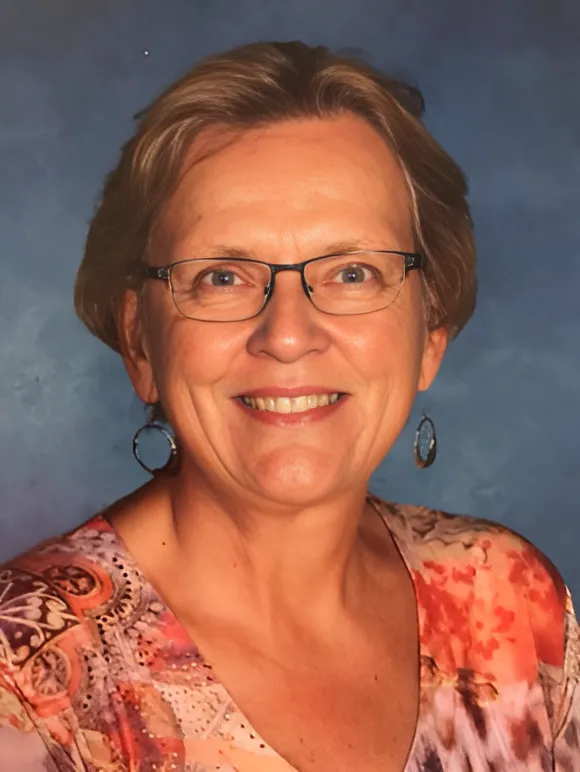 Portrait of Susan Espiritu—a woman with tan skin, short dark blonde hair—wearing a floral blouse and smiling gently toward the camera against a blue background.