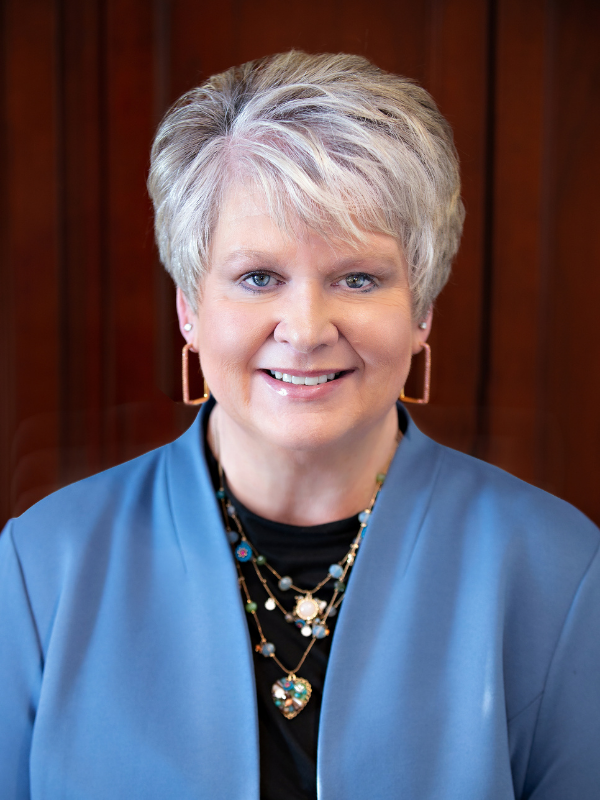 Headshot of a light-skinned woman with short gray hair, wearing a blue jacket and layered necklaces, smiling in front of a warm-toned wooden background.