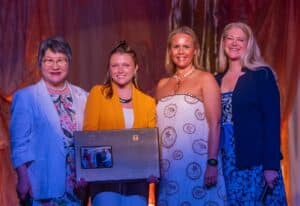 Four women stand on a stage wearing semi-formal wear. One is holding a large plaque.